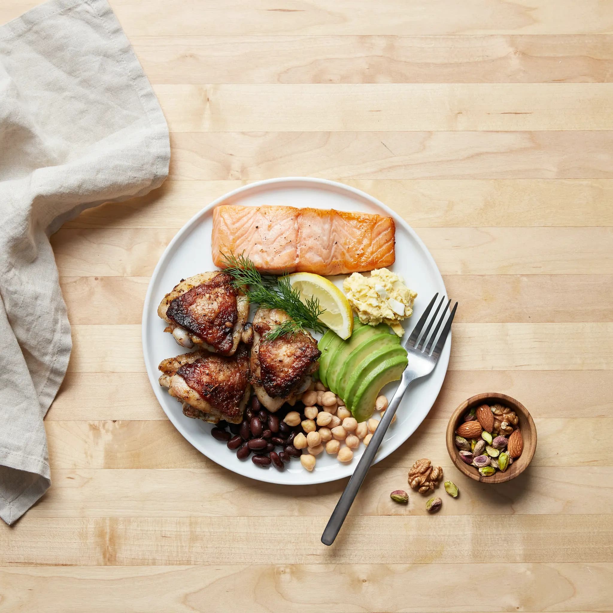 Plate of salmon, roasted chicken, avocado, chickpeas and black beans with a bowl of nuts